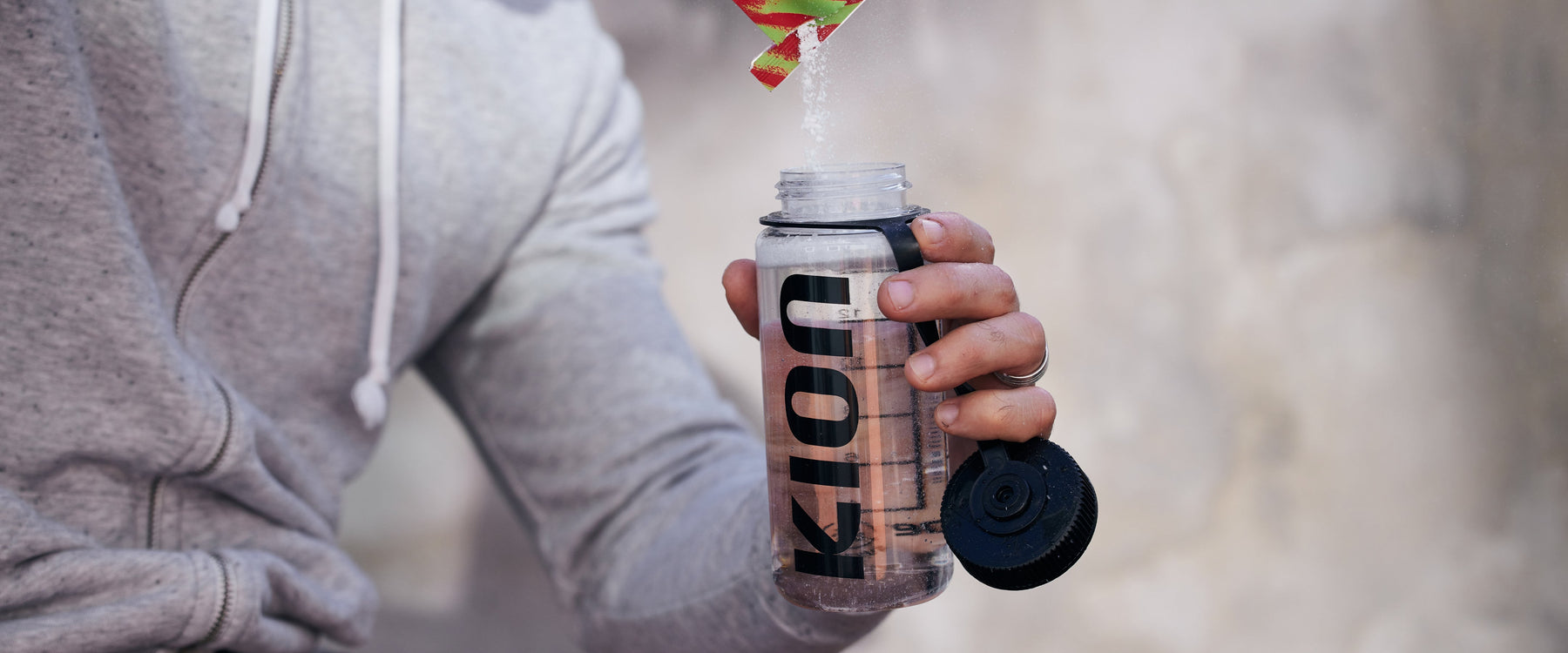 A person in a gray hoodie holds a clear Kion shaker bottle with a black cap and a green-and-red straw, with liquid being poured into it against a blurred beige background.