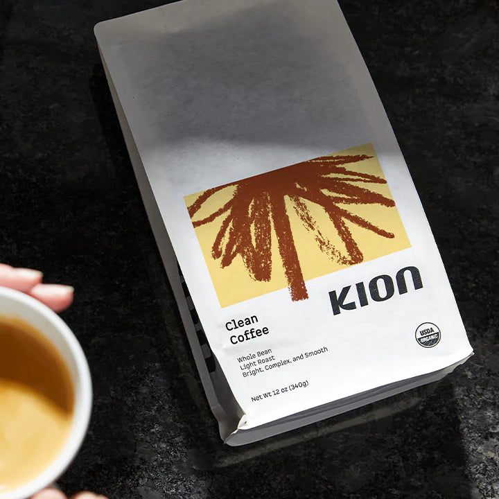 A bag of KION Clean Coffee whole bean light roast organic coffee sits on a dark countertop, with a hand holding a cup of coffee partially visible on the left.