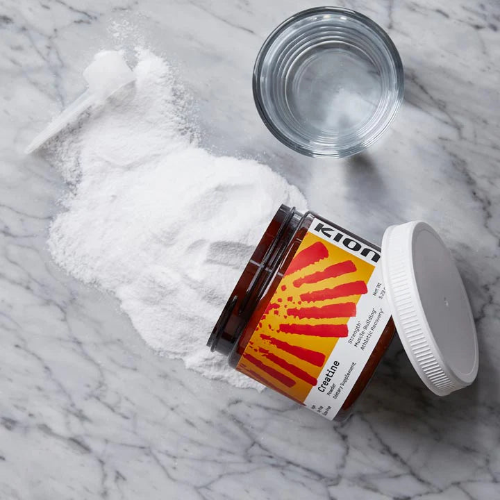 An open amber jar labeled Kion Creatine with white powder spilled beside it, a white screw cap nearby, and a small glass dish on a light marble surface.