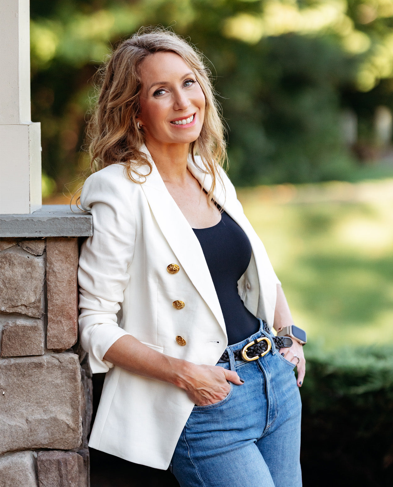 A woman posing outdoors in a white double-breasted blazer with gold buttons, a black top, and blue jeans, leaning against a stone pillar.