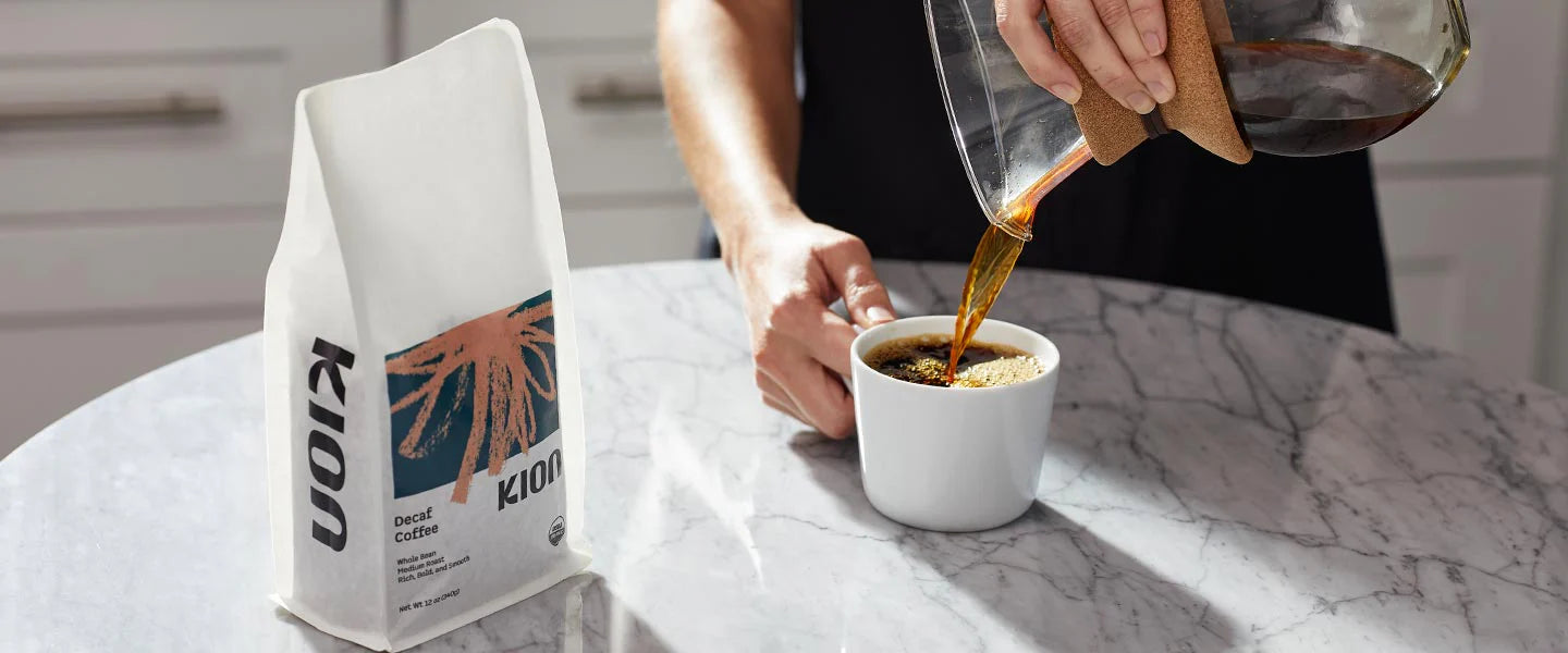 A white bag of Kion coffee stands on a marble countertop next to a person pouring hot coffee from a glass carafe into a white mug.