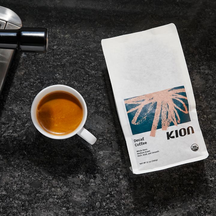 A white bag of Kion Decaf Coffee lies on a dark speckled countertop next to a white cup of espresso, with part of a coffee maker visible at the top left.