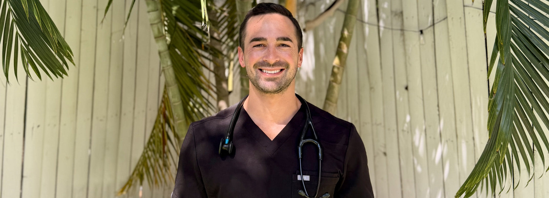 Man in black medical scrubs with a stethoscope standing against a light wooden panel background with green leaves.