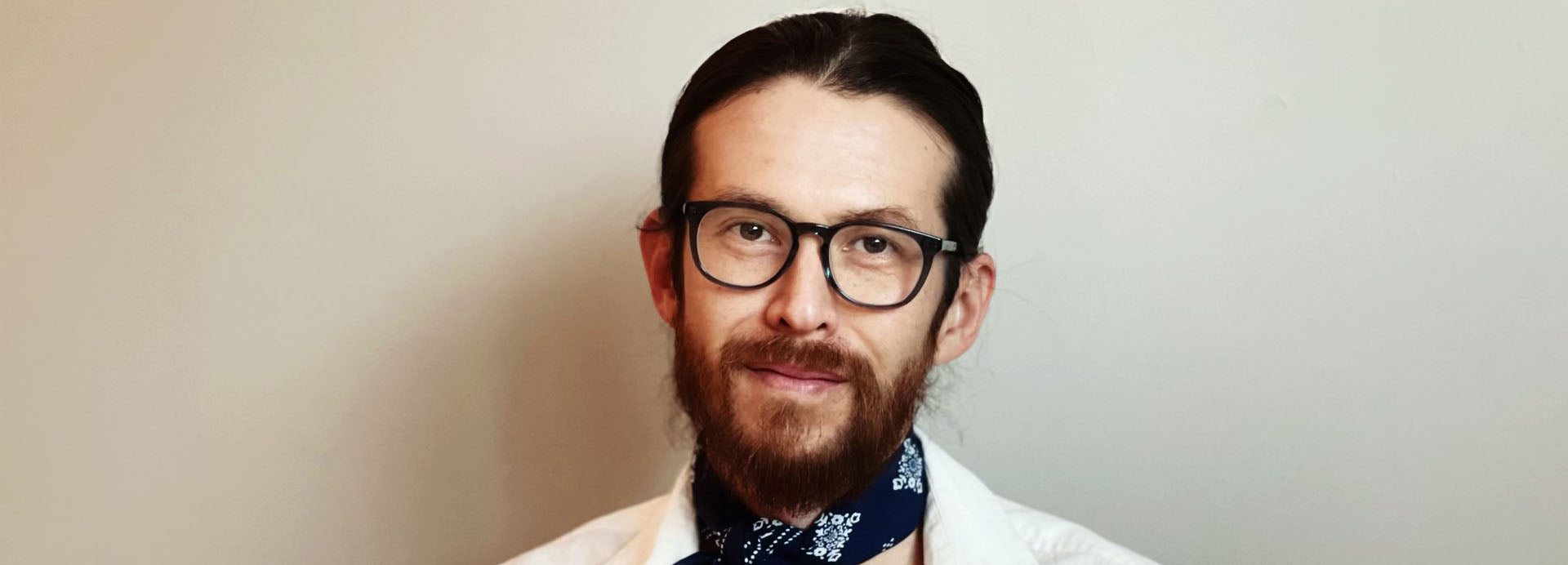 Man with glasses and a beard wearing a patterned scarf against a plain background