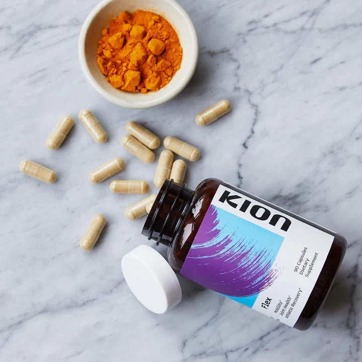 An open bottle of Kion Flex joint supplement lies on a white marble surface with scattered beige capsules and a small bowl of bright orange powder nearby.