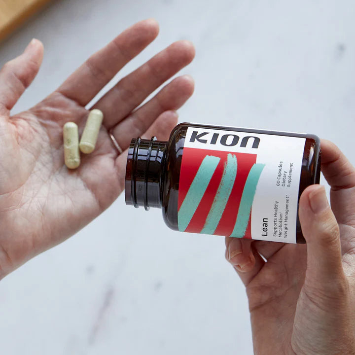 A person holds an amber supplement bottle labeled KION Lean while showing two beige capsules in their other hand against a light marble background.