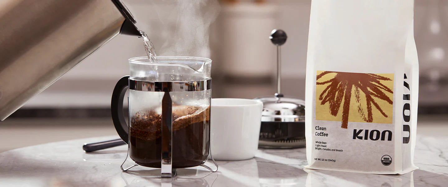 A bag of Kion Clean Coffee stands on a marble countertop beside a glass coffee maker with steaming coffee, a white mug, and a metal coffee press in a softly blurred kitchen setting.
